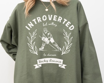 Puede incluir: Sudadera verde con texto blanco que dice "Introvertido pero dispuesto a hablar de romance de hockey". La sudadera presenta un gráfico de un jugador de hockey.