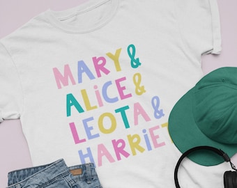 May include: A white t-shirt with the text "MARY & ALICE & LEOTA & HARRIE" printed in colorful letters. The shirt is laid out on a pink surface with a pair of blue denim shorts, a green baseball cap, black headphones, and a wooden skateboard.