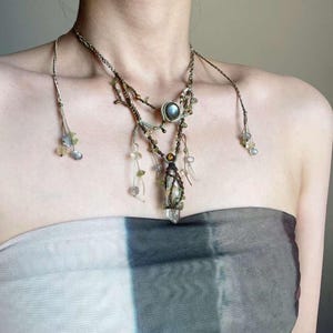 May include: A layered necklace featuring a crystal pendant, brown and green beads, and a blue stone. The necklace is crafted from natural materials and has a bohemian aesthetic.