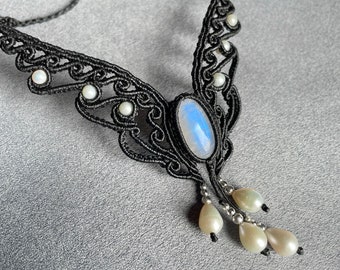 May include: A black macrame necklace with a large oval moonstone pendant and pearl accents. The necklace has a delicate, intricate design and is perfect for a bohemian or ethereal style.