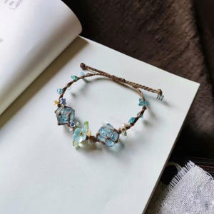 May include: A delicate bracelet made with brown cord, blue and green crystals, and small flower accents.