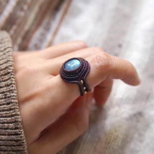 Puede incluir: Un anillo con una piedra preciosa azul engastada en una banda tejida de cordón morado oscuro. El anillo está en un dedo.