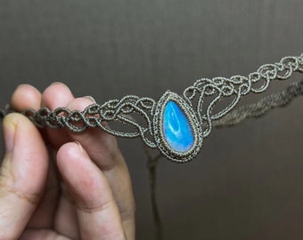 Könnte beinhalten: Ein handgefertigtes Makramee-Stirnband mit einem zentralen, tropfenförmigen blauen Edelstein. Das Stirnband ist aus einem gedämpften graubraunen Kordel gefertigt und weist komplizierte Knotenmuster auf. Der Edelstein hat eine leuchtend blaue Farbe.