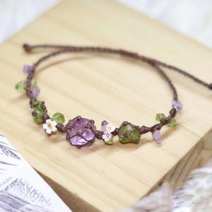May include: A delicate bracelet made with brown cord, amethyst, green peridot, and white flower accents. The bracelet is tied with a knot and features a cluster of amethyst crystals.