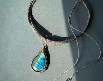 Puede incluir: Un collar de macramé marrón con un colgante de piedra de labradorita en forma de lágrima. El collar tiene dos borlas largas con pequeñas cuentas blancas y azules.