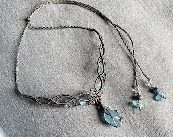 May include: A delicate silver necklace with a large, faceted blue gemstone pendant. The necklace is made of wire and features a woven design. The pendant is surrounded by smaller blue gemstones.