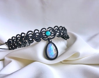 May include: A black macrame choker necklace with a silver bead detail and a teardrop-shaped iridescent gemstone pendant. The pendant is accented with a smaller turquoise gemstone.