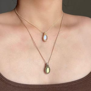 May include: Two layered necklaces with oval-shaped pendants. The top pendant has a light blue stone, while the bottom pendant has a yellow-green stone. Both pendants are encased in a woven, beige-colored setting and hang from delicate chains.