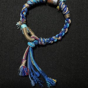 May include: A handmade bracelet featuring a braided design with blue, yellow, and red threads. The bracelet has a tassel of blue and red threads. The bracelet is on a black background.