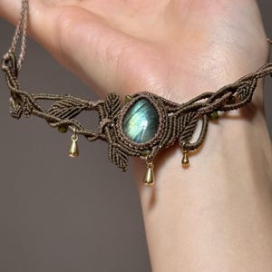 May include: A handcrafted macrame necklace featuring a central oval labradorite stone with iridescent blue and green hues. The brown cord design includes leaf-like details and small gold-colored teardrop charms, creating a bohemian aesthetic.
