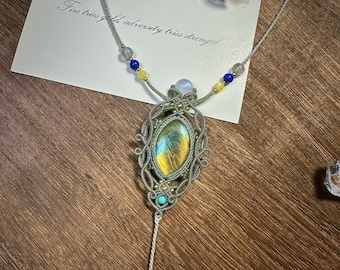 May include: A silver necklace with a green and gold gemstone pendant. The pendant is surrounded by a silver wire design. The necklace is strung with white, blue, and yellow beads. The text "Fire tries gold, adversity tries strength" is visible in the background.