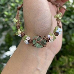 Peut inclure: Bracelet en macramé fait à la main avec une pierre verte centrale, entourée de petites perles vertes et blanches, et de minuscules fleurs bleues et blanches. Le bracelet est en cordon marron.