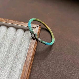 May include: A braided bracelet with a gradient of blue, green, and yellow hues. The bracelet features a silver-colored clasp with a decorative design. The bracelet is displayed on a wooden jewelry tray with a gray fabric interior.