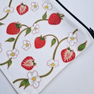May include: White canvas pouch with a black zipper, adorned with a repeating pattern of red strawberries, white blossoms, and green leaves. The design features whole and sliced strawberries, creating a fresh, summery aesthetic.
