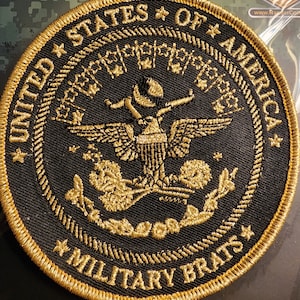 May include: A black and gold embroidered patch with the words "United States of America" and "Military Brats" around a golden eagle with a diamond on its chest.