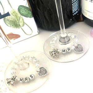 May include: Two wine glasses with decorative charms. One charm spells "MRS" and the other "MR". Each charm features a silver bell, a heart, and beads. The background includes a wine bottle and a floral design.