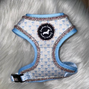 Może przedstawiać: Jasno niebieski szelki dla psa z białym tłem i wzorem niebieskich kokardek. Szelki mają różowe i srebrne cyrkonie wokół krawędzi i czarną klamrę. Szelki mają okrągłą naszywkę z tekstem "Lunabears Customs" i sylwetką psa rasy dachshund.