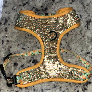 Puede incluir: Un arnés para perro amarillo con un patrón floral y un diseño brillante. El arnés tiene un anillo en D de metal y un cierre de hebilla.