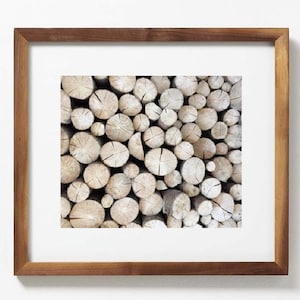 May include: A framed print featuring a close-up of a stack of cut logs. The logs are light brown with visible growth rings and cracks. The frame is a dark brown wood.