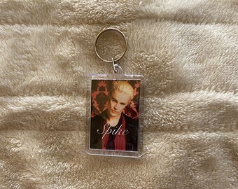 May include: A clear, rectangular keyring with a portrait of a man with blonde hair and the name "Spike" in cursive. The portrait has a dark red and black patterned background. The keyring has a gold-coloured ring.