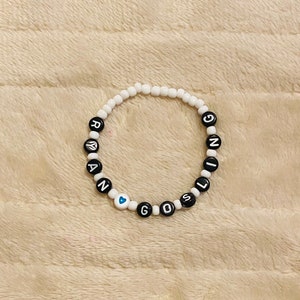 May include: White beaded bracelet with black letter beads spelling "RANGING GIRL". The bracelet includes a blue heart bead. The bracelet is on a beige, textured surface.