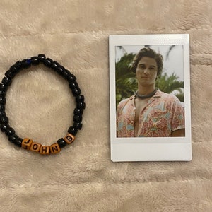 May include: A black beaded bracelet with the name "JOHN B" spelled out in wooden letter beads. Next to the bracelet is a photo of a man wearing a patterned shirt.