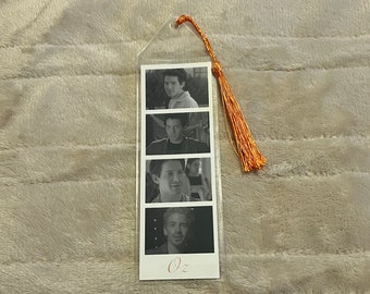 Puede incluir: Marcapáginas transparente con cuatro imágenes en blanco y negro de una persona. Tiene una borla naranja en la parte superior. La palabra "Oz" está impresa en naranja claro en la parte inferior del marcapáginas.