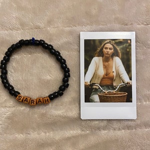 May include: A black beaded bracelet with the name "SARAH" spelled out in wooden letter beads. Next to the bracelet is a polaroid-style photo of a person riding a bicycle.