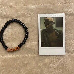 May include: A black beaded bracelet with the word "POPE" spelled out in wooden letter beads. Next to the bracelet is a Polaroid-style photo of a man wearing a hat.