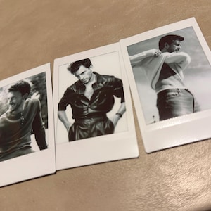 May include: Three black and white Polaroid-style photographs featuring a man in various poses. The photos show the man in different outfits, including a leather shirt and a sweater. The images are arranged on a neutral-toned surface.