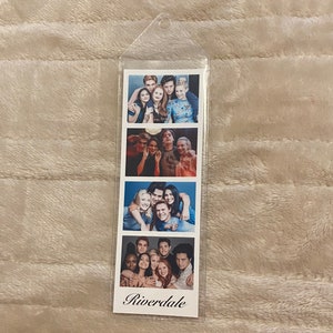 May include: A clear plastic bookmark featuring four photo strips with images of a cast from the television show "Riverdale." The bottom strip has the word "Riverdale" printed in a script font.
