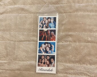 May include: A clear plastic bookmark featuring four photo strips with images of a cast from the television show "Riverdale." The bottom strip has the word "Riverdale" printed in a script font.
