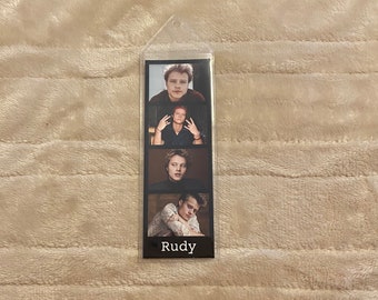 Puede incluir: Marcapáginas de plástico transparente con cuatro imágenes de una persona. El nombre "Rudy" está impreso en blanco en la parte inferior. El fondo es de color beige claro.
