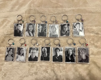 May include: A selection of rectangular keyrings displaying black and white portraits. Each keyring has a silver ring and a small, coloured gem. The keyrings are presented on a soft, textured surface.