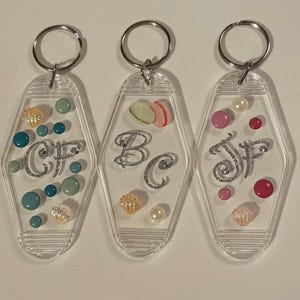 May include: Three clear acrylic keyrings with silver rings. Each keyring features a unique design with initials, decorative beads, and seashells. The initials are in a silver glitter font. The beads are in shades of teal, pink, and pearl.