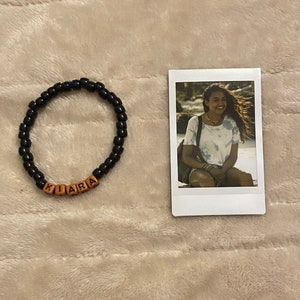 May include: A black beaded bracelet with the name "KIARA" spelled out in wooden letter beads. Next to the bracelet is a small photograph of a woman wearing a white t-shirt.