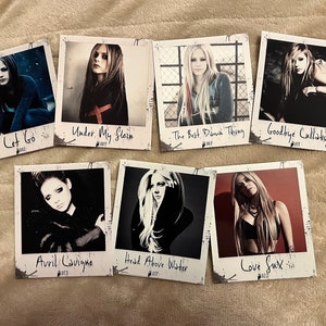 Könnte beinhalten: Sieben quadratische Drucke mit Bildern von Avril Lavigne, jeweils mit einem anderen Albumtitel: "Let Go", "Under My Skin", "The Best Damn Thing", "Goodbye Lullaby", "Avril Lavigne", "Head Above Water" und "Love Sux". Die Drucke haben einen Polaroid-Stil mit weißen Rändern und Text.