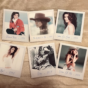 Peut inclure: Six tirages de style polaroid présentant des images de Shania Twain de différentes années. Les tirages incluent le texte "Shania Twain ~ 1993", "The Woman In Me ~ 1995", "Come On Over ~ 1997", "Up! ~ 2002", "Now ~ 2017" et "Queen Of Me ~ 2025". Les tirages sont disposés sur une surface beige.