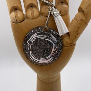 May include: A silver astronaut helmet keychain with a black glitter background and a white tassel.