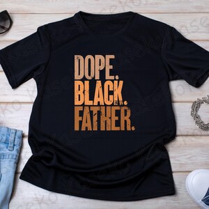 May include: Black t-shirt with the words "DOPE. BLACK. FATHER." in an orange, distressed font. The shirt is displayed on a light wood surface, with sunglasses, jeans, and a sneaker visible.