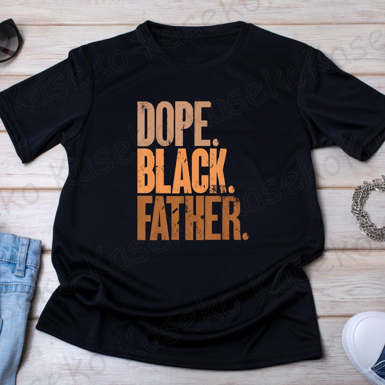 May include: Black t-shirt with the words "DOPE. BLACK. FATHER." in an orange, distressed font. The shirt is displayed on a light wood surface, with sunglasses, jeans, and a sneaker visible.