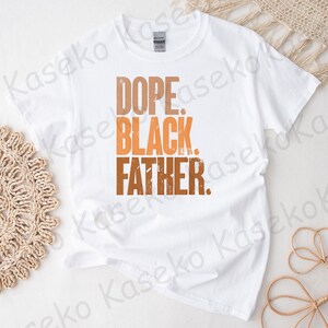 May include: White t-shirt with the words "DOPE. BLACK. FATHER." in a gradient of brown and orange. The text has a distressed, textured appearance. The shirt is made of a soft material.