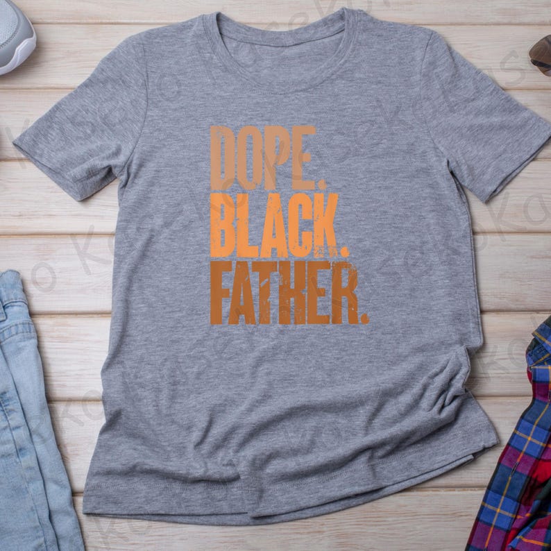 May include: A gray t-shirt with the words "DOPE. BLACK. FATHER." in orange and brown block letters. The shirt is made of a soft material and has short sleeves. The shirt is laying on a light wood surface.
