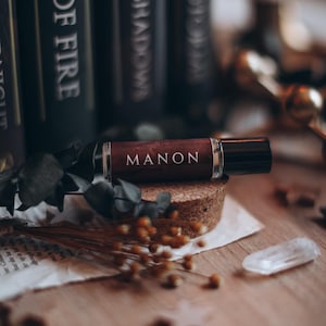May include: A dark brown perfume bottle labelled "MANON" sits on a cork base. Dried flowers, a clear crystal, and books with white text titles surround the bottle. The background is blurred, creating a warm, inviting aesthetic.