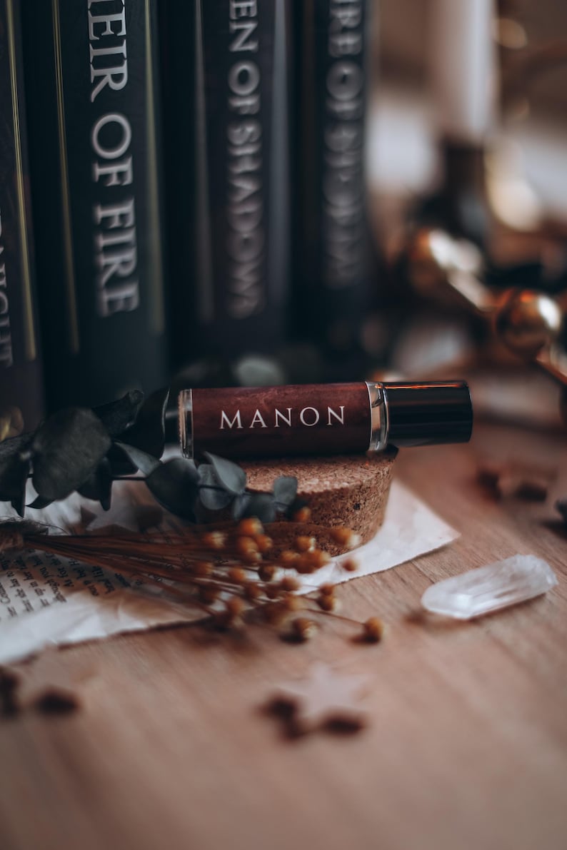 May include: A dark brown perfume bottle labelled "MANON" sits on a cork base. Dried flowers, a clear crystal, and books with white text titles surround the bottle. The background is blurred, creating a warm, inviting aesthetic.