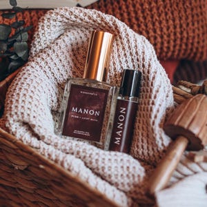 May include: Two perfume bottles with gold lids and the word "MANON" printed on them, in a woven basket with a beige knitted blanket. The larger bottle has the text "PLUM + LIGHT MUTE". A wooden mushroom decorates the scene.