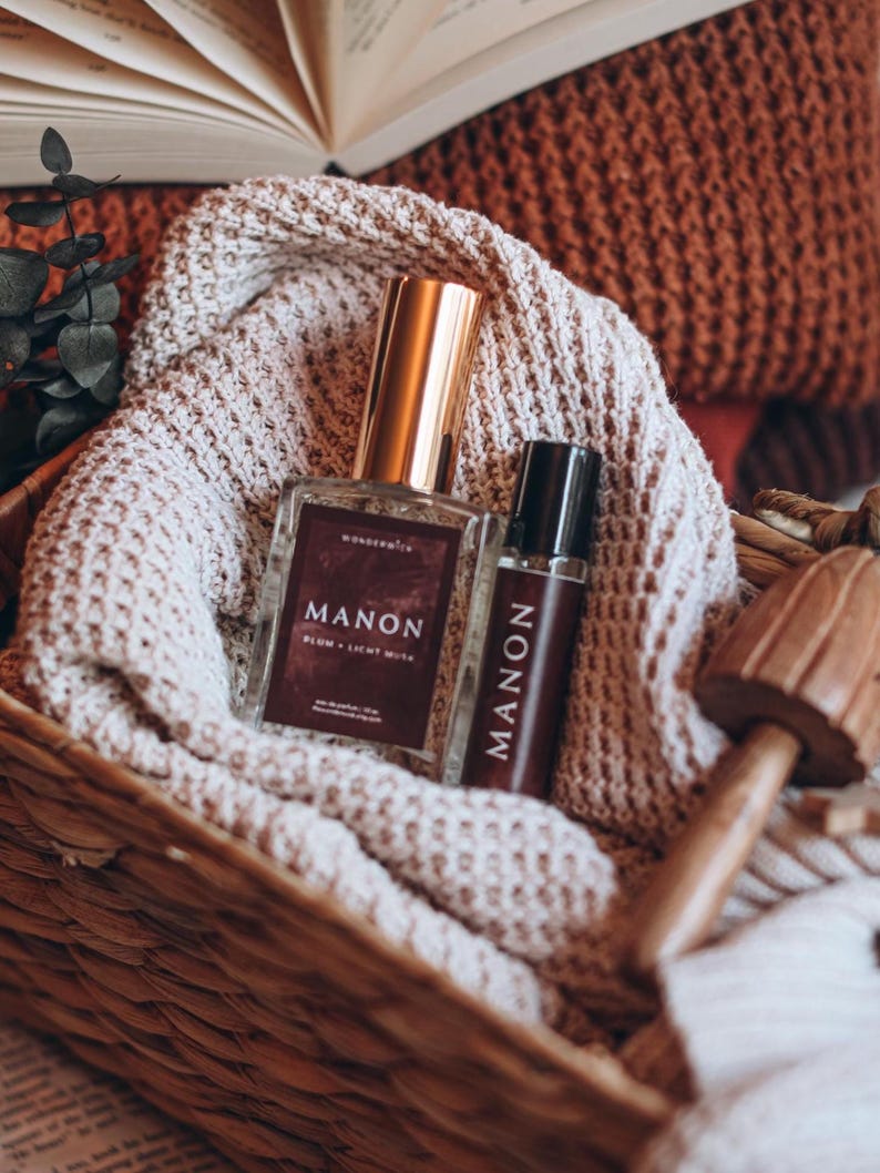 May include: Two perfume bottles with gold lids and the word "MANON" printed on them, in a woven basket with a beige knitted blanket. The larger bottle has the text "PLUM + LIGHT MUTE". A wooden mushroom decorates the scene.