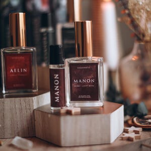 May include: Three perfume bottles with gold lids and rectangular labels. The labels read "AELIN", "MANON", and "MANON Plum + Light Musk". The bottles are displayed on wooden blocks, with books and candles in the background.