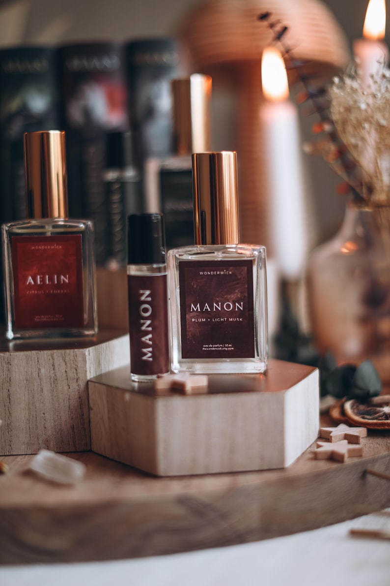 May include: Three perfume bottles with gold lids and rectangular labels. The labels read "AELIN", "MANON", and "MANON Plum + Light Musk". The bottles are displayed on wooden blocks, with books and candles in the background.