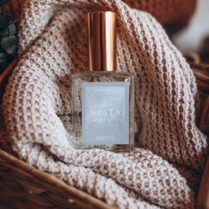 May include: A rectangular glass perfume bottle with a gold cap, labeled "NESTA LEATHER & STEEL", rests on a beige knitted blanket inside a woven basket. The bottle contains 50 ml of perfume. The background includes a book and more knitted items.
